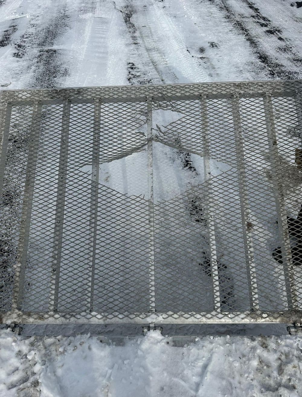 Metal grating gate covered in snow on a winter road, showcasing a textured surface.