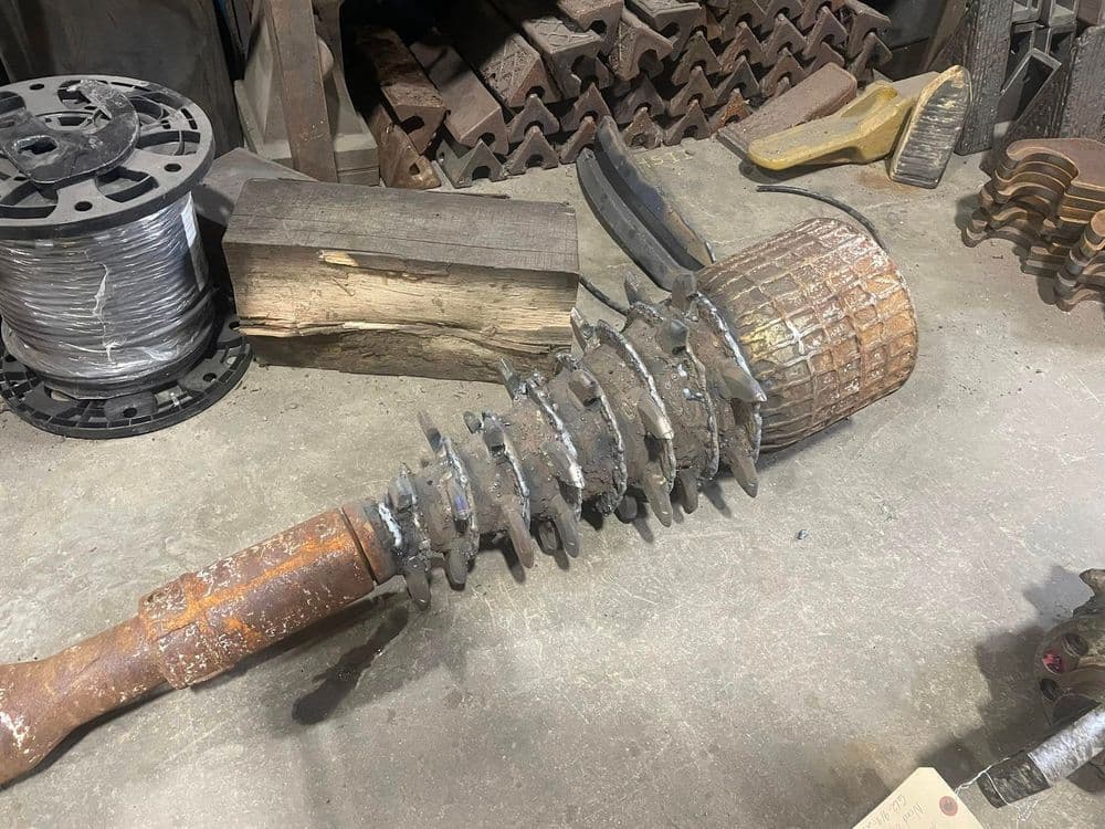 Rusty metal auger drill resting on workshop floor with industrial tools in background.